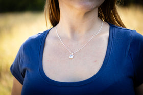 Person wearing a blue shirt with a necklace featuring a small square cremation pendant in a natural setting.