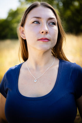 Person wearing a blue shirt with a necklace featuring a small square cremation pendant in a natural setting.