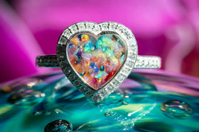 Bedazzled Heart Cremation Ring with rainbow selection of crushed opal. The ring is sitting atop a glass surface, with the stone facing the viewer. Sterling Silver Ring for Ash, Sterling Silver Memorial Jewelry, Remembrance Jewelry, Silver Ring
