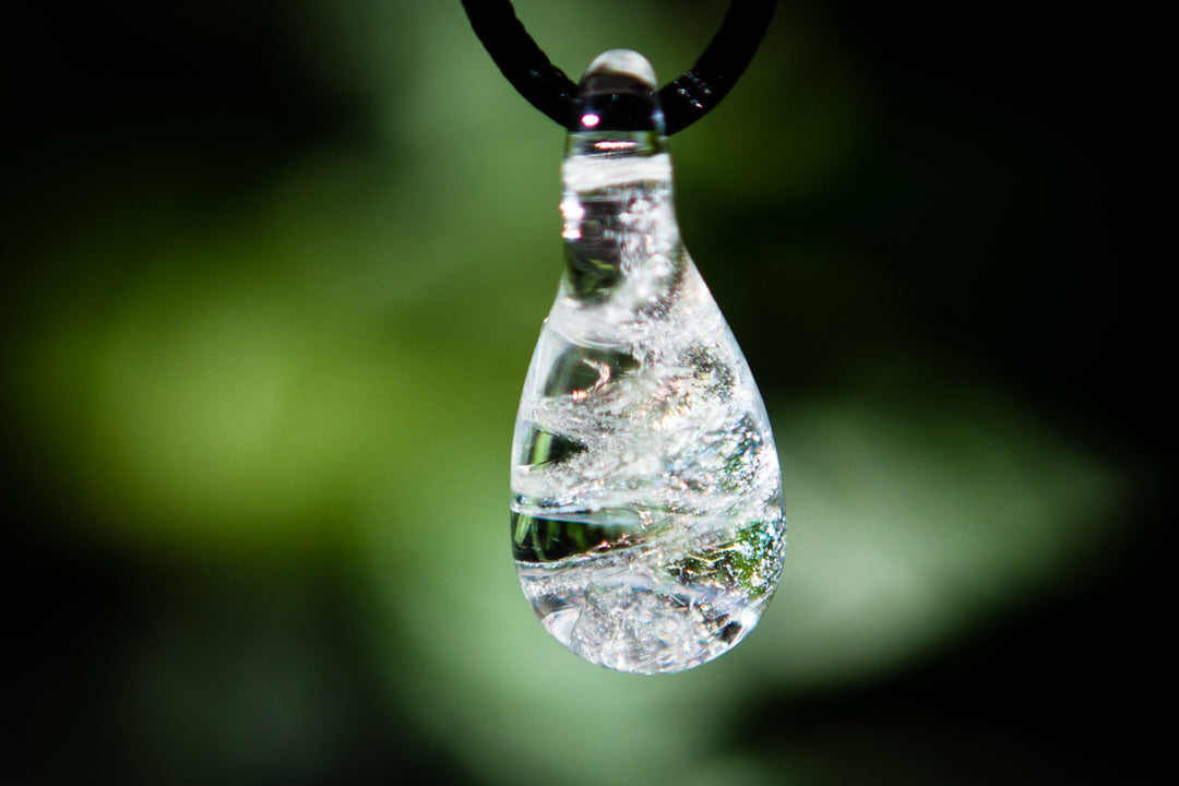 Teardrop glass pendant filled with clear liquid, suspended by a black cord against a blurred green background