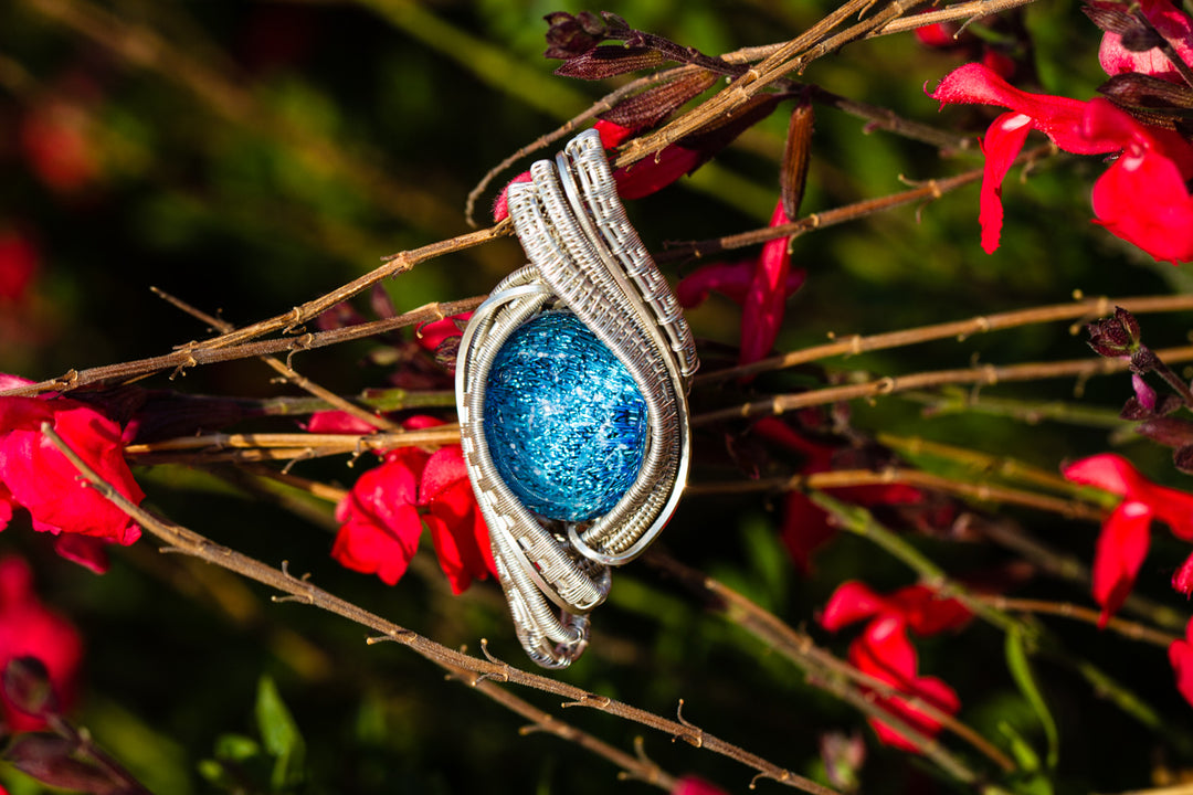 Blue glittery oval cremation stone wrapped in silver wire pendant resting on red flowering branches.