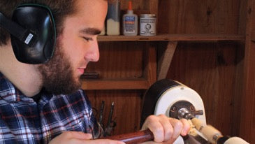 Bearded man wearing headphones operates a wood lathe, turning a wooden piece in a workshop.