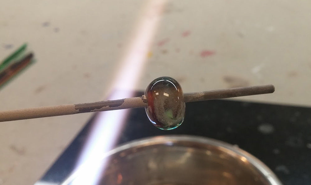 Small glass bead on a rod above a torch flame in a glass studio during beadmaking.