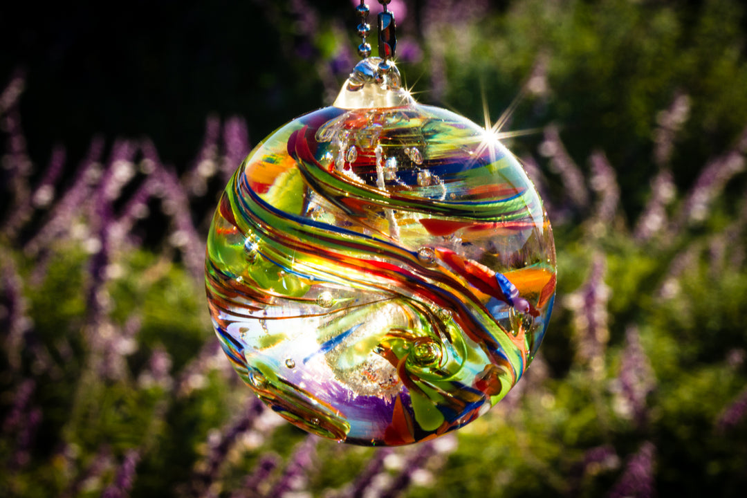 Colorful glass cremation ornament with swirling ribbons hanging outdoors against a blurred garden background.