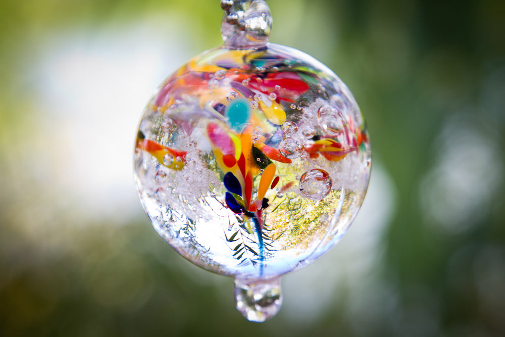 Clear glass cremation orb pendant filled with multicolored glass pieces and plant motifs, hanging outdoors.