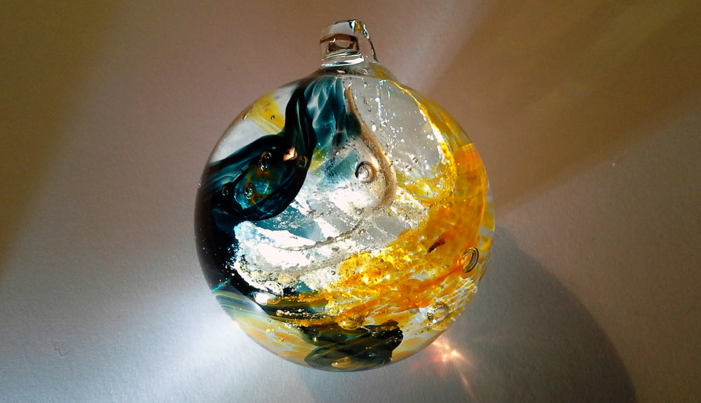 Glass cremation orb with teal and orange swirls and white accents resting on a pale surface.