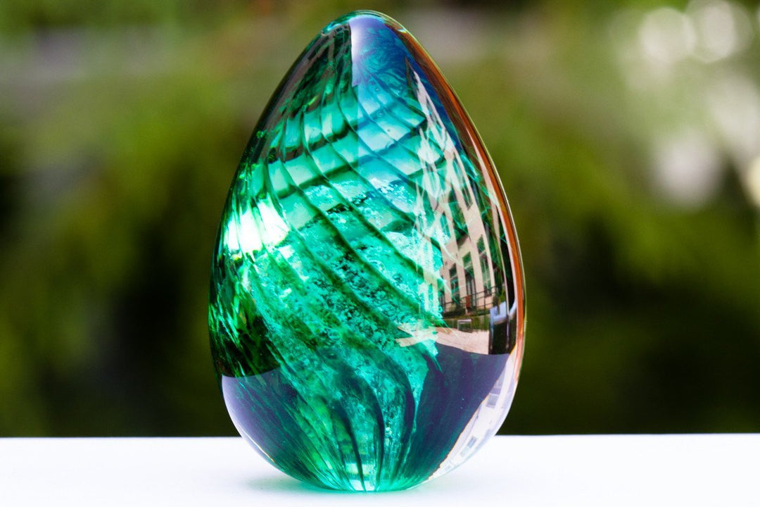 Teardrop-shaped green cremation glass ornament reflecting a building, placed on a white surface.