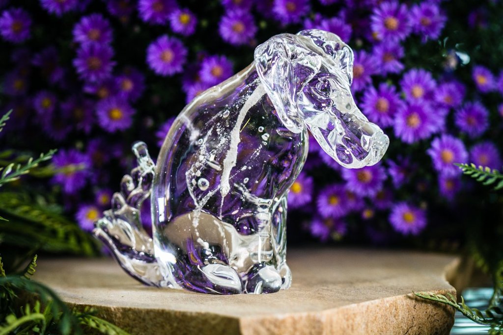Clear cremation glass dog sculpture on a wooden surface with vibrant purple flowers in the background.