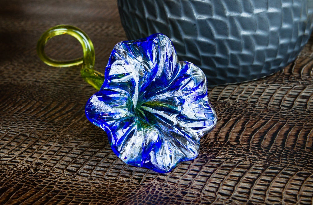 Blue glass flower with green stem resting on a textured brown surface beside a gray patterned vase.