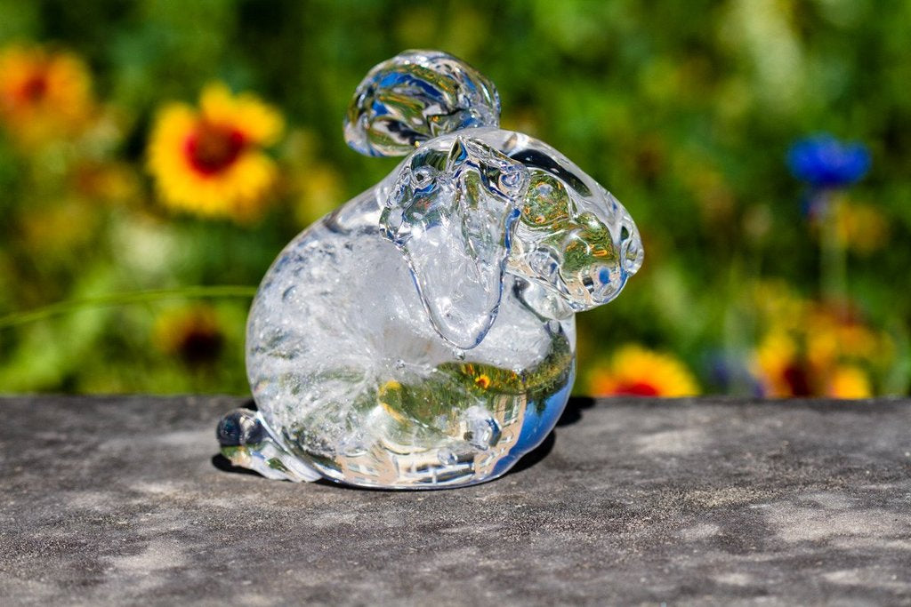 Crystal cremation glass rabbit sculpture resting on a stone surface with a blurred garden of colorful flowers in the background
