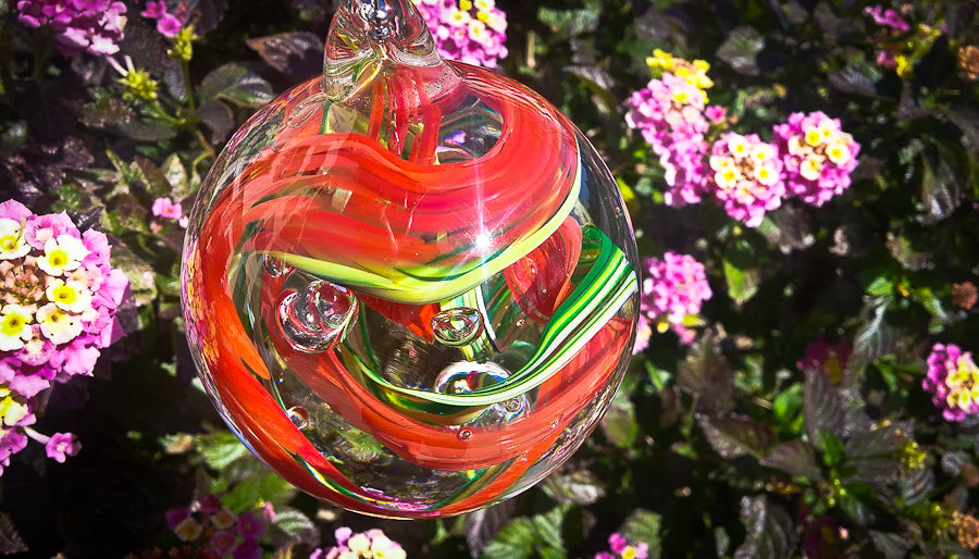 Colorful glass memorial ornament with red and green ribbons swirling inside, set against purple blossoms.