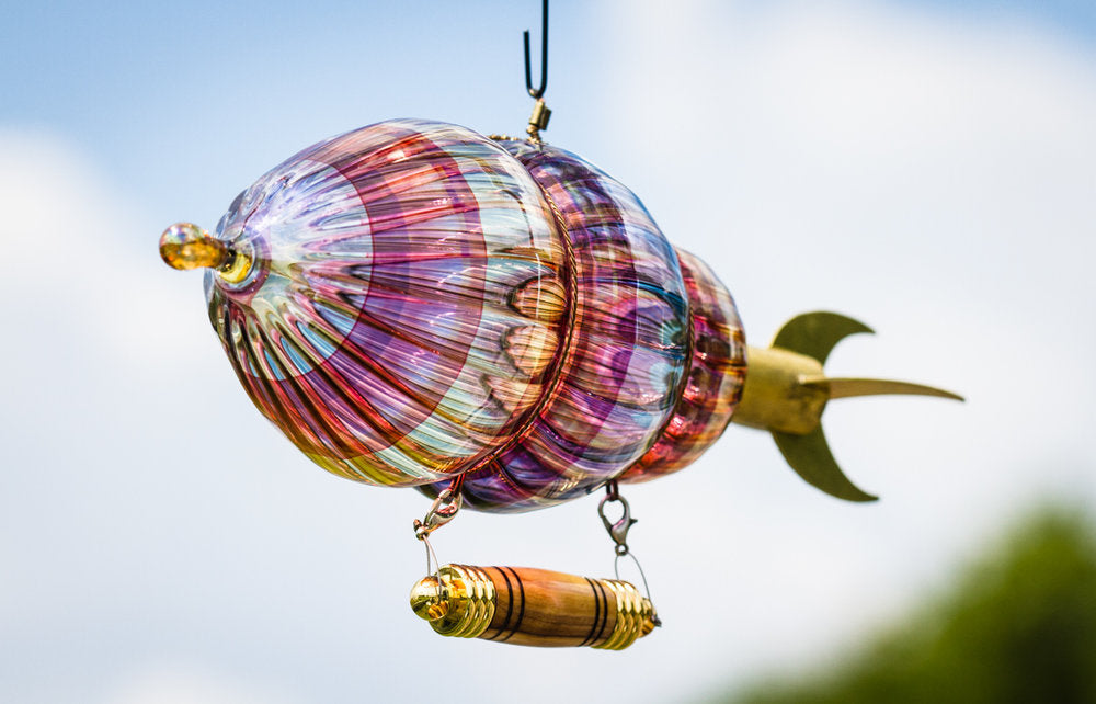 Colorful glass airship sculpture hanging in the air with a wooden gondola against a blue sky.
