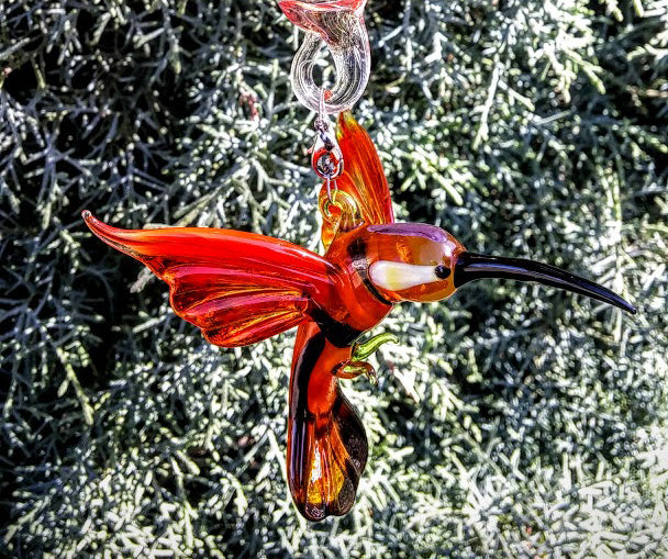 Red glass hummingbird embellishment with a clear hanger attached to a memorial cremation ash orb against evergreen background.