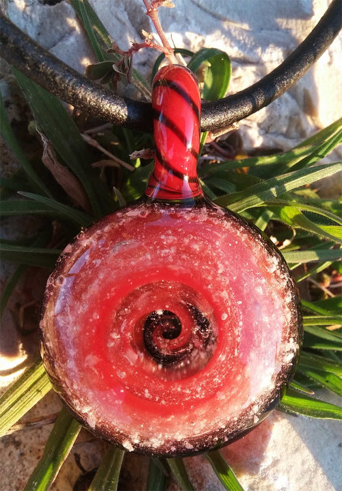 Red swirl glass keepsake pendant resting on rocks beside green plants.