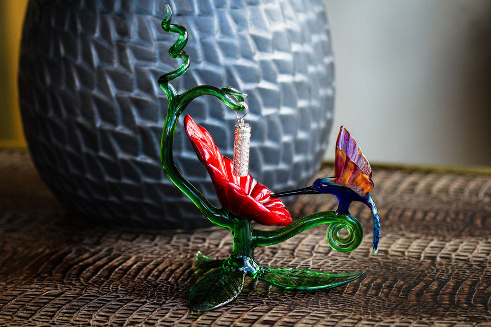 Colorful glass hummingbird sculpture perched on a green twisting stem beside a red bloom.