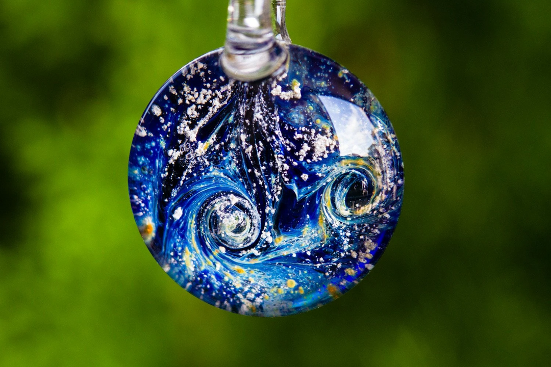 Blue marbled cremation glass pendant hanging from a cord against a green blurred background