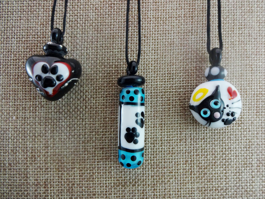 Three pet themed glass pendants on cords a paw print heart, a blue polka dot paw print tube, and a round cat face charm.