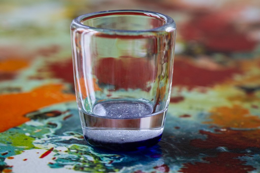 Clear cremation glass shot glass on a vibrant paint splatter tabletop, a memorial keepsake