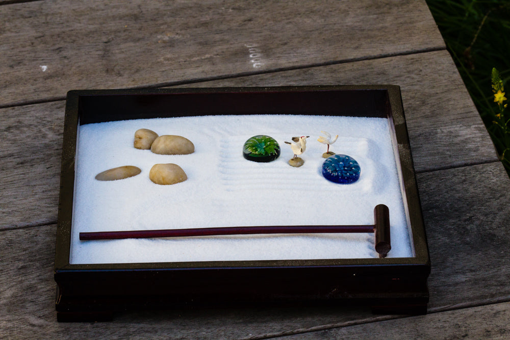 Zen garden tray with white sand left stones a green glass pebble a blue pebble two tiny bird figurines and a red wooden rake.