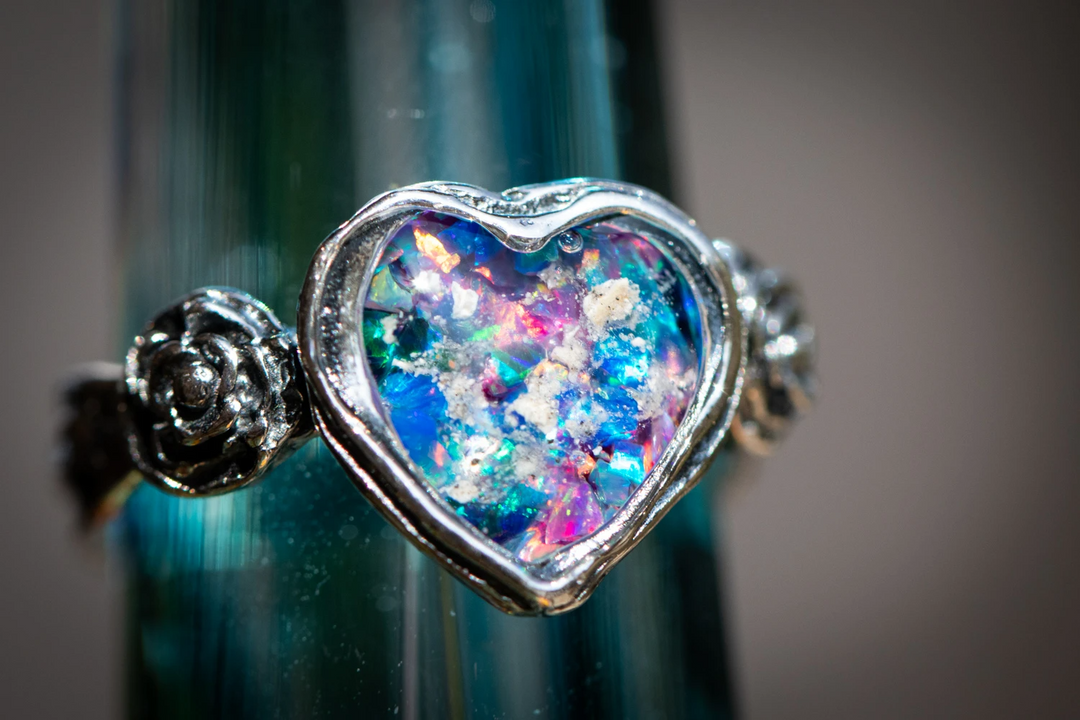 Heart shaped silver cremation pendant filled with iridescent colorful material, flanked by decorative silver rosette ends.