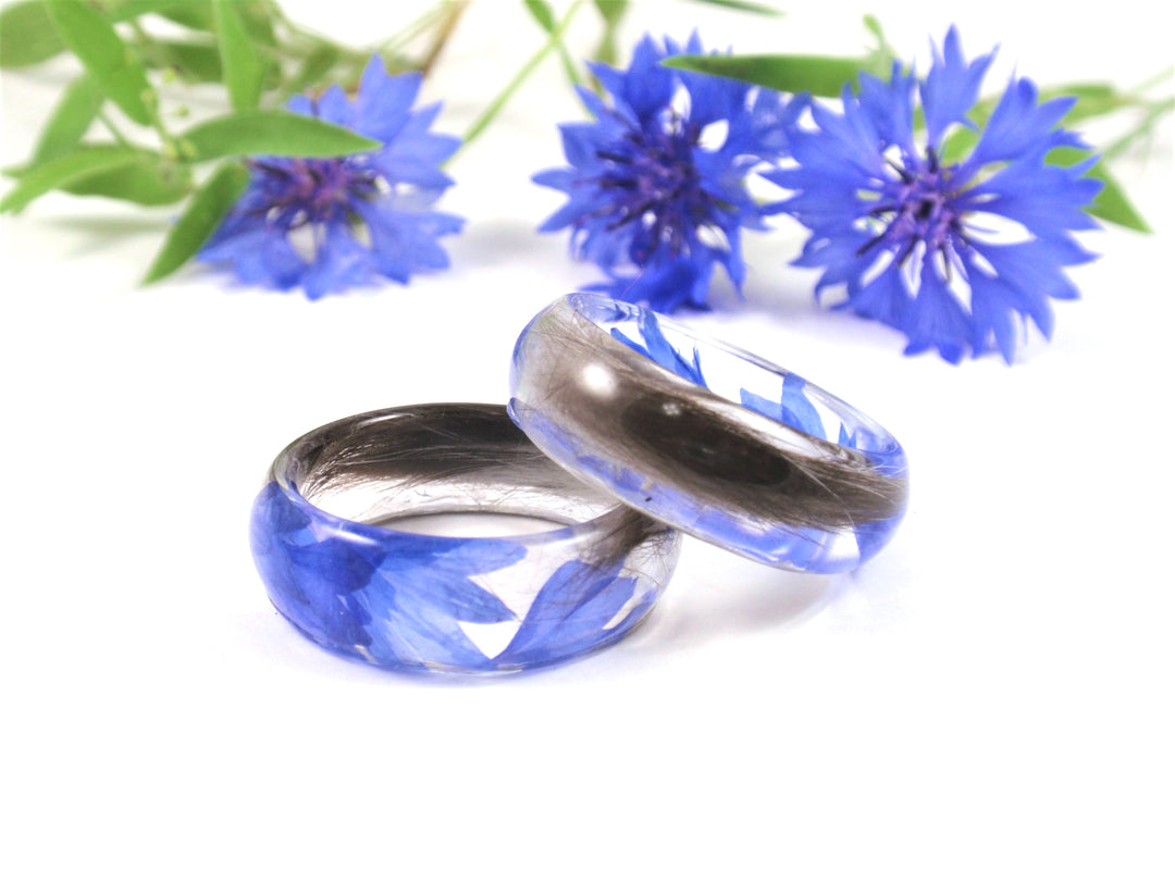 Blue crystal resin cremation rings with silver interiors resting on a white background with blue flowers in the background