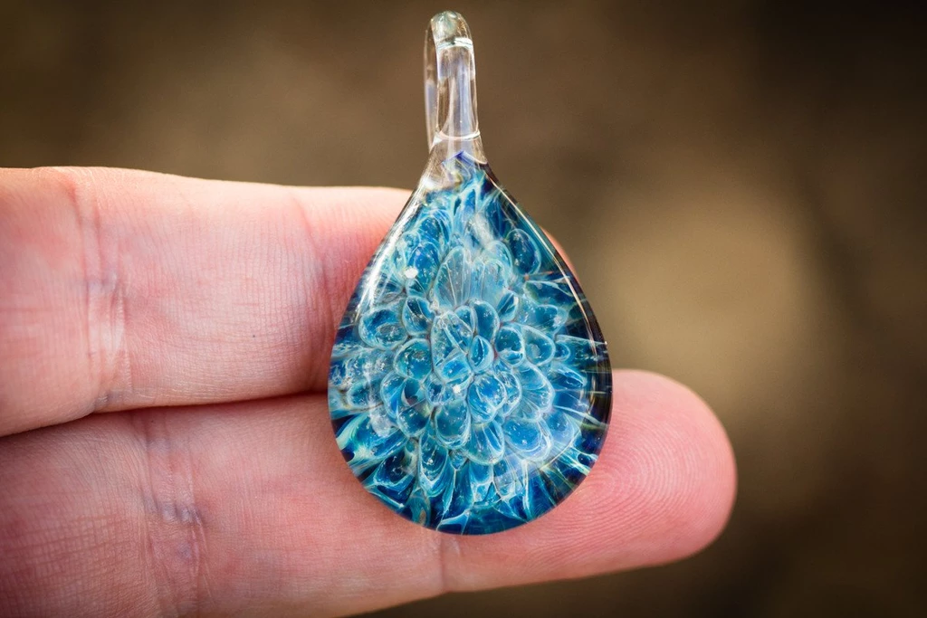 Blue teardrop glass pendant with an intricate flower pattern held between fingers.