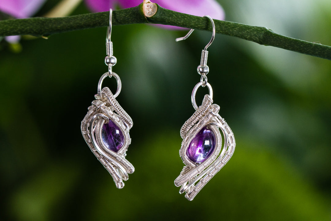 Wire wrapped silver earrings with purple gemstones hanging against a blurred green background