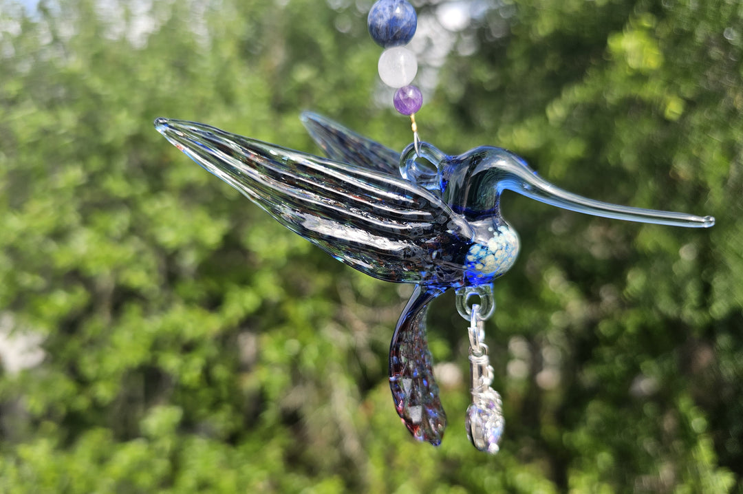 Profile view of blue glass hummingbird pendant with beaded strand and crystal leaf charm against green foliage.