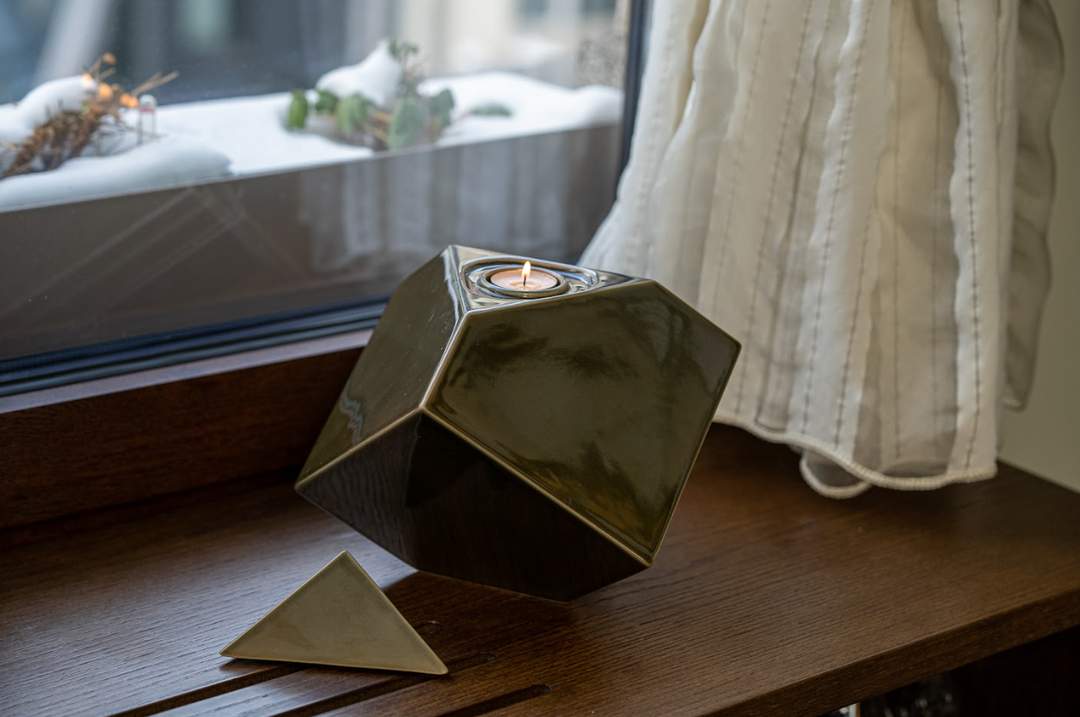 Abstract Ceramic Urn in dark glaze perched on edge with a lit candle on top and a detached triangular cap on a wooden sill by a window.