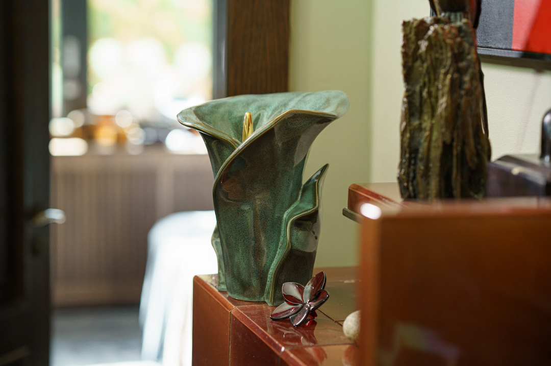 Turquoise Calla Lily ceramic urn resting on a wooden cabinet in a bright room with a decorative metal flower nearby.
