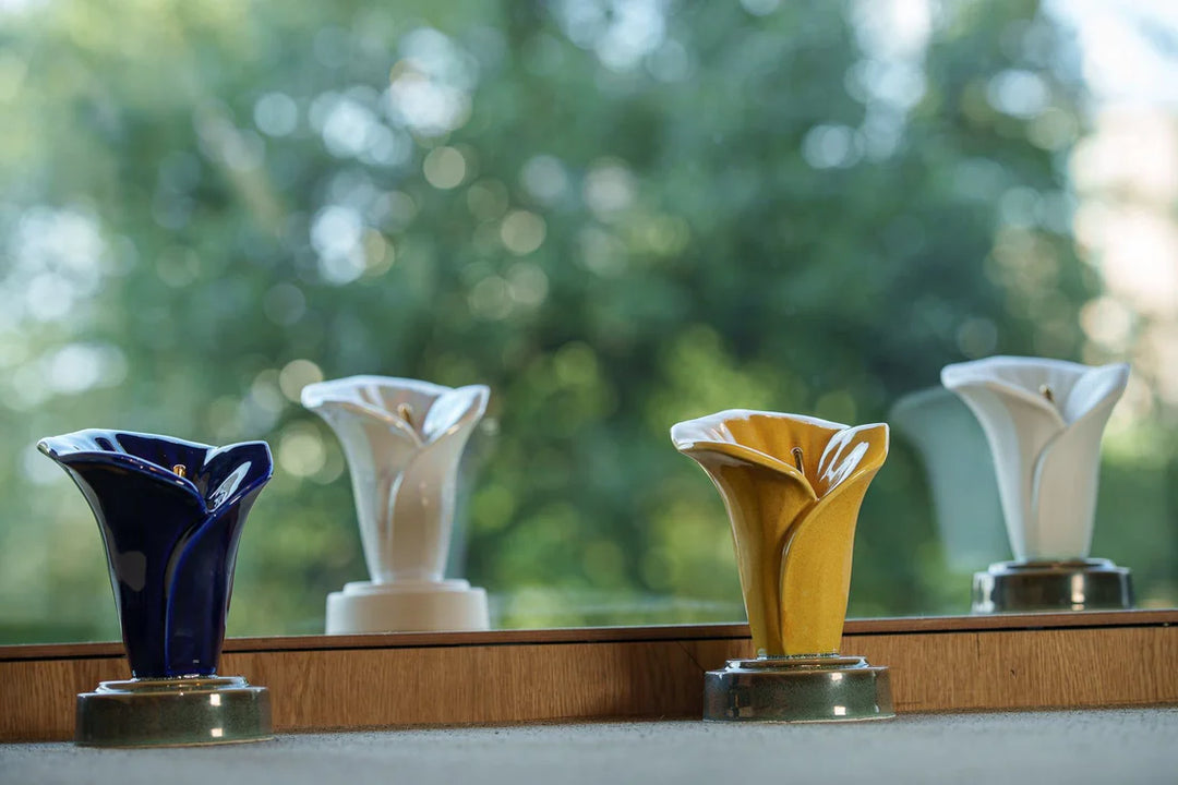 Four colorful Calla Lily Keepsake Ceramic Urns blue white and gold arranged on a wooden ledge with a blurred greenery background.