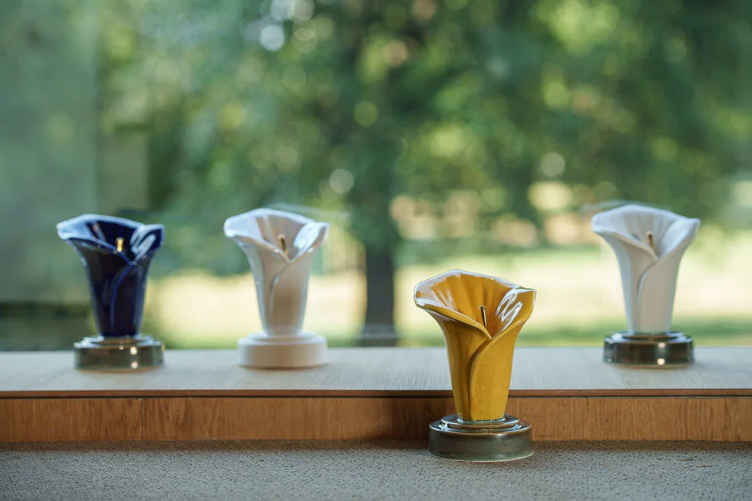 Set of four calla lily keepsake ceramic urns in blue white and gold tones displayed on pedestals along a sunlit windowsill.