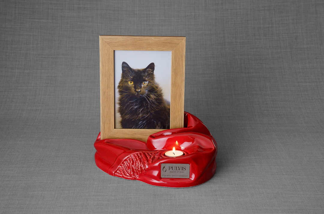 Cat portrait in a wooden frame on a glossy red sculpted ceramic urn with a candle.