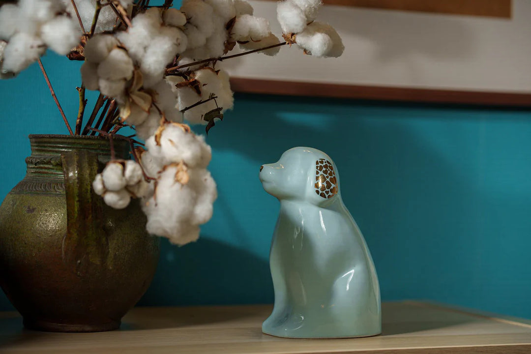 Light blue dog shaped ceramic urn with lattice cutout on the ear sits on a shelf beside a green vase with cotton stems