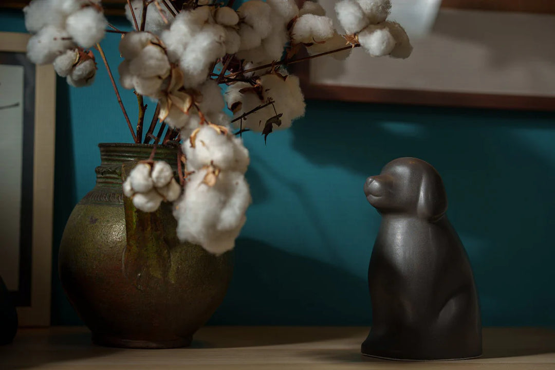 Cuddle Dog ceramic urn in a dark glaze sits on a wooden surface beside a rustic vase with cotton stems.