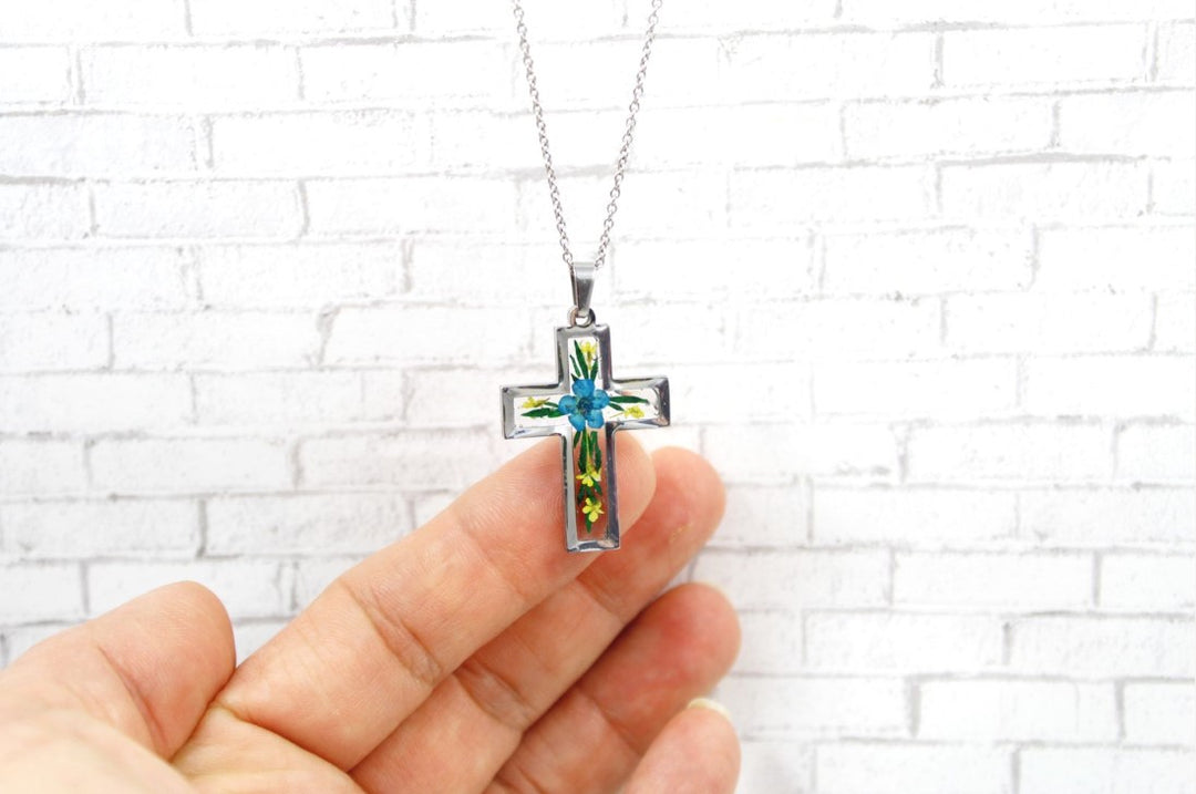 Close up of the crystal resin cross pendant featuring a blue flower center and green leaves, hung on a silver chain and held by fingers against a white brick background.