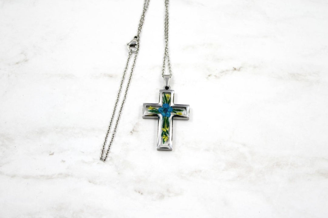 Crystal resin cross pendant with blue flower and green leaves inside a silver frame on a long silver chain against a white brick backdrop.