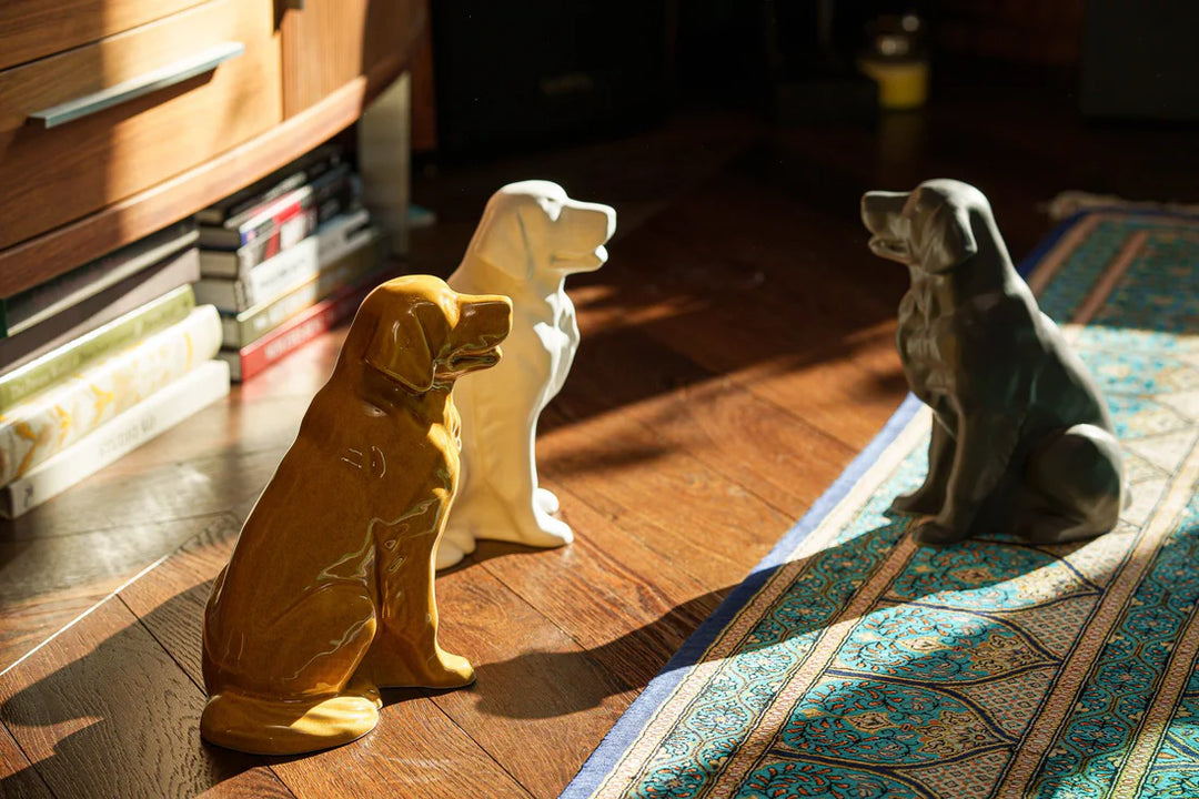 Three ceramic dog figurines in brown, white, and gray sit on a wooden floor beside a patterned rug.