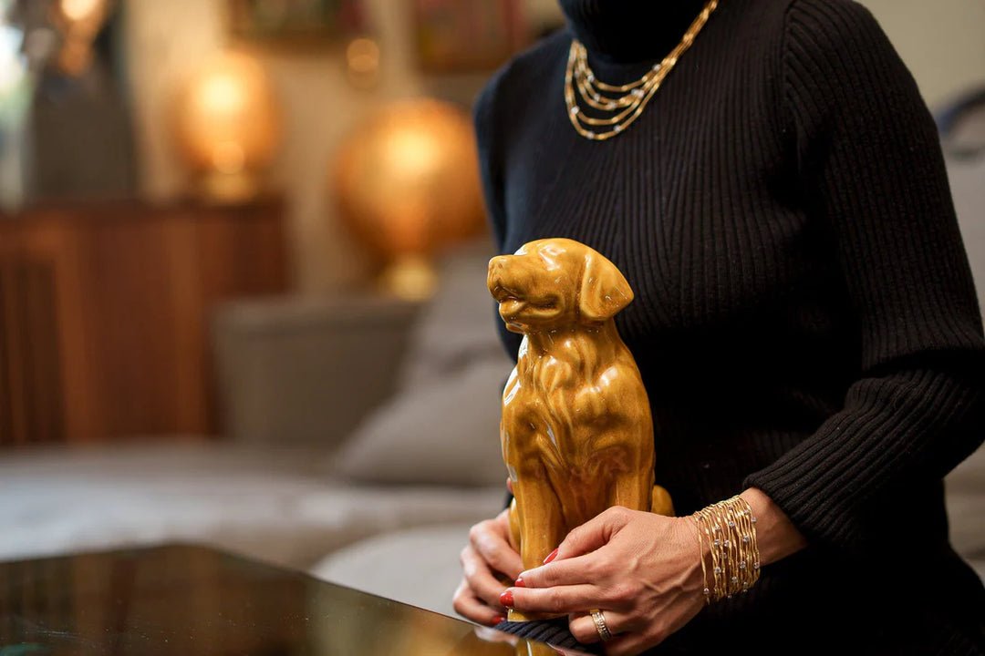Person in a black knit sweater with gold bracelets holds a glossy golden retriever ceramic figurine on a coffee table.