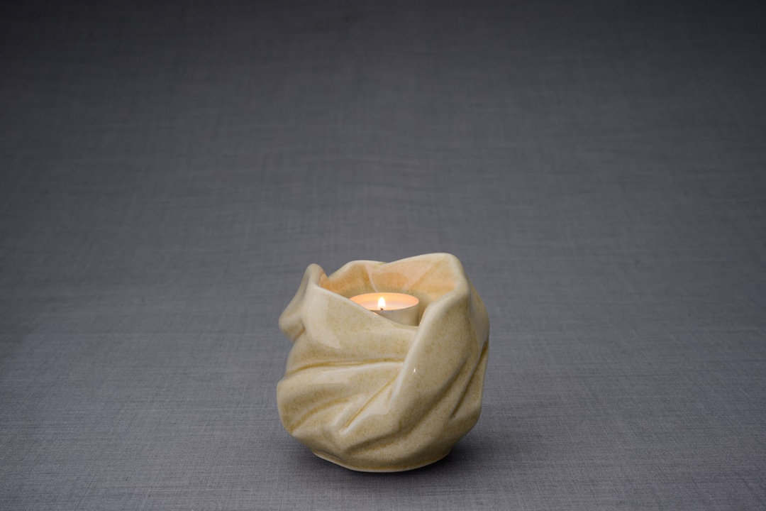 The Holy Mother beige glaze ceramic urn with soft sculpted folds and a lit tealight candle.