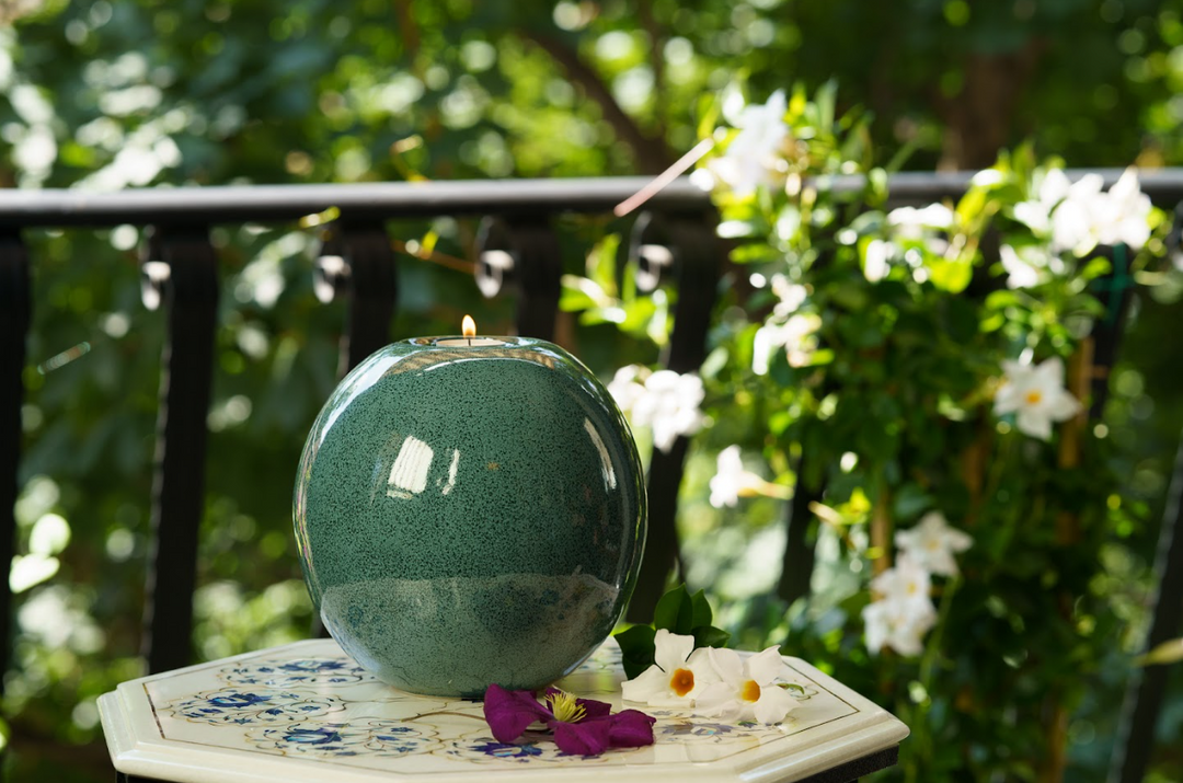 Teal Harmony ceramic urn with candle on top sits on an outdoor table surrounded by white and purple flowers and greenery.