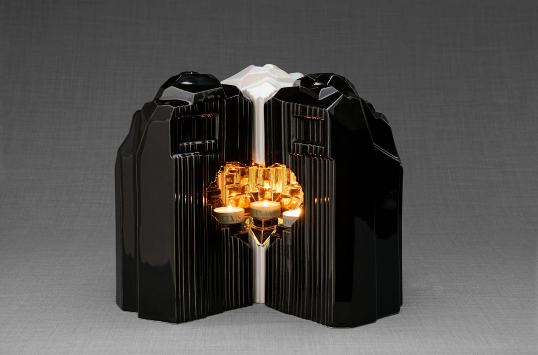 Black glossy heart shaped ceramic urn halves opened to reveal a candle holder with lit candles.