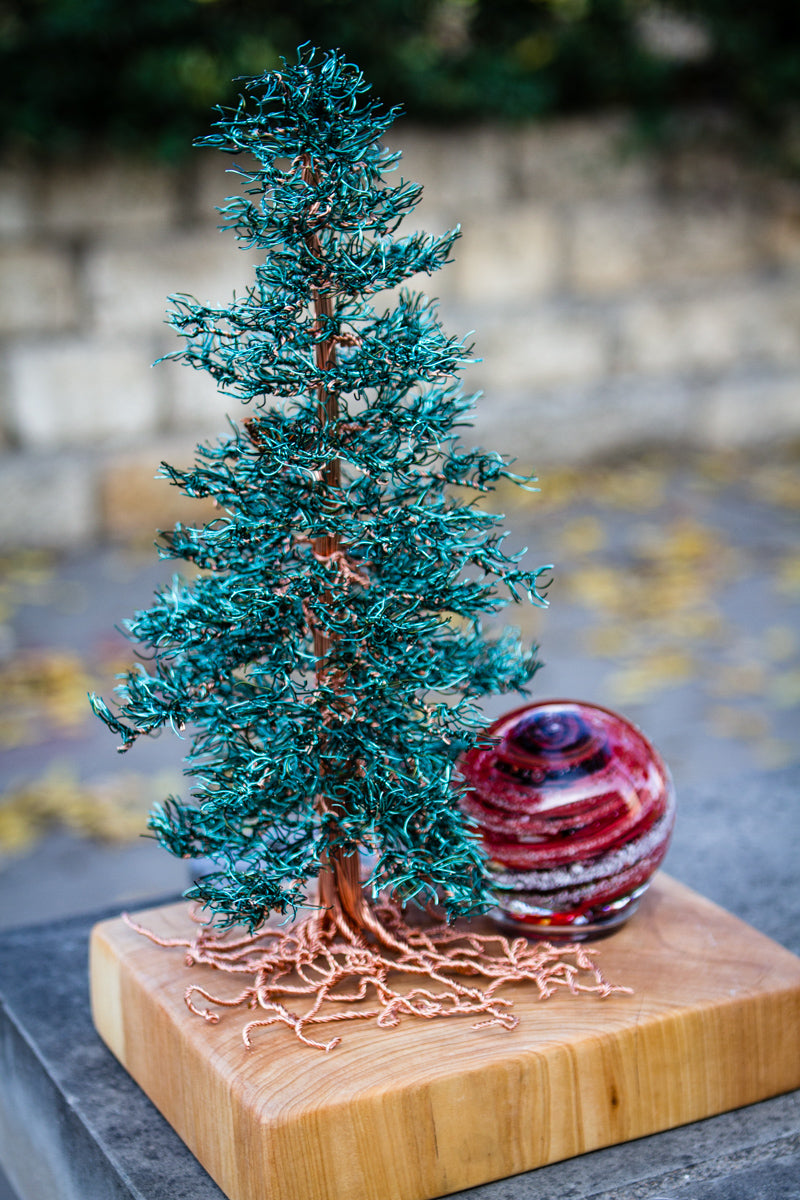Teal Spruce Tree of Life mounted on a wooden base with copper roots and a red glass orb beside it outdoors on a stone surface.