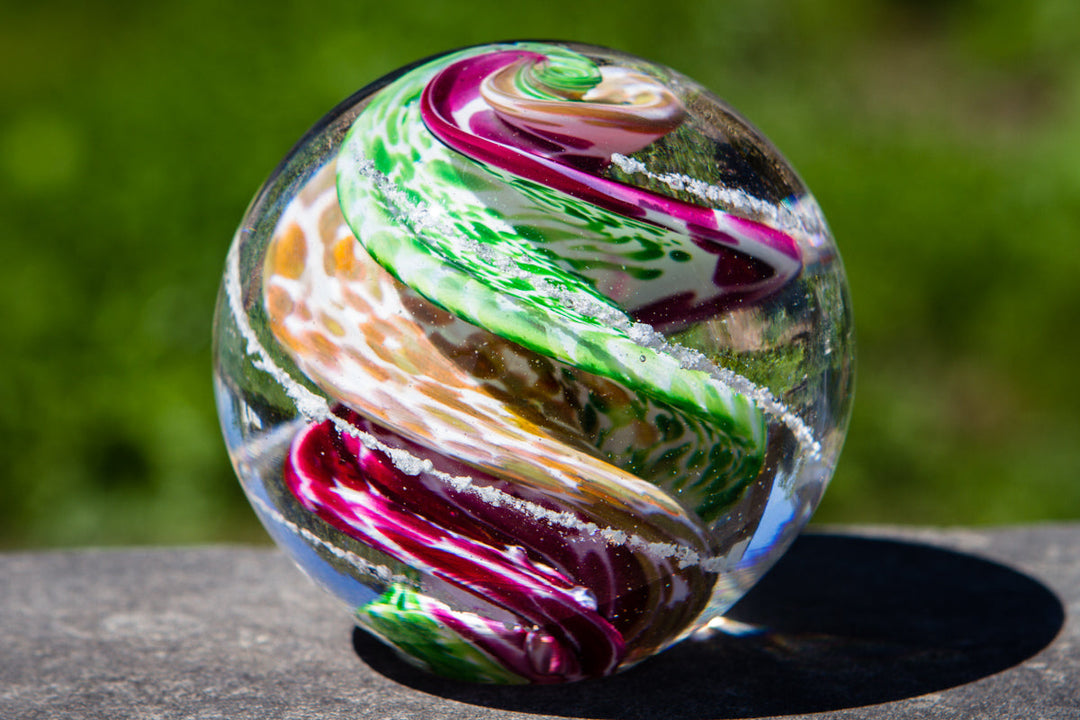Swirled multicolor glass cremation orb with cremains resting on a dark outdoor surface