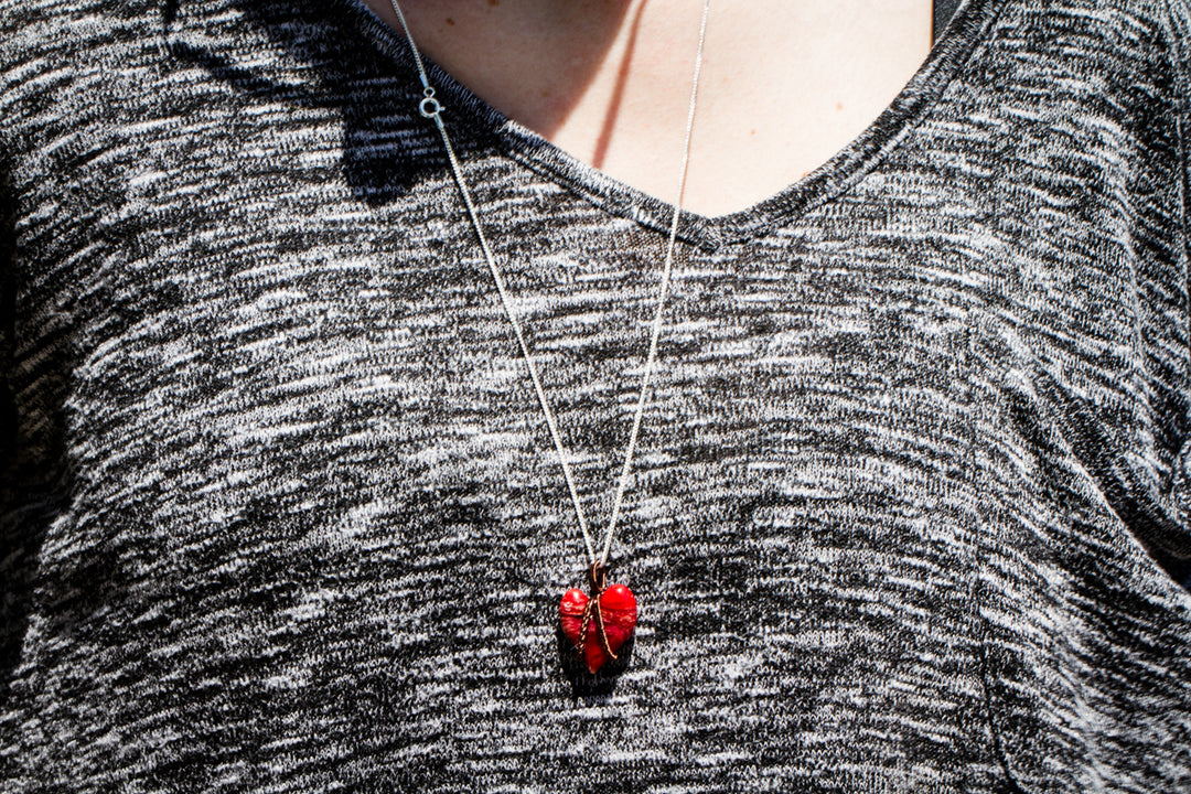 Red glass heart cremation ashes pendant wrapped in silver wire on a silver chain against a gray knit top.