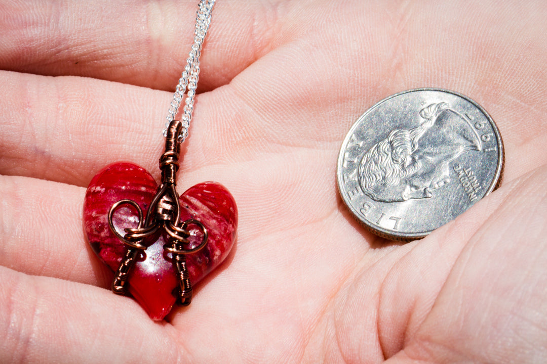 Red glass heart pendant wrapped in copper wire on a silver chain beside a quarter for scale