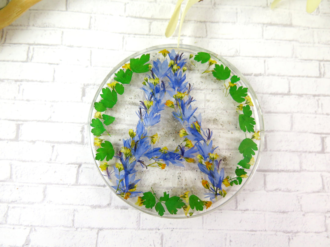 Round clear memorial coaster with blue flower arrangement forming a letter A and green leaves.