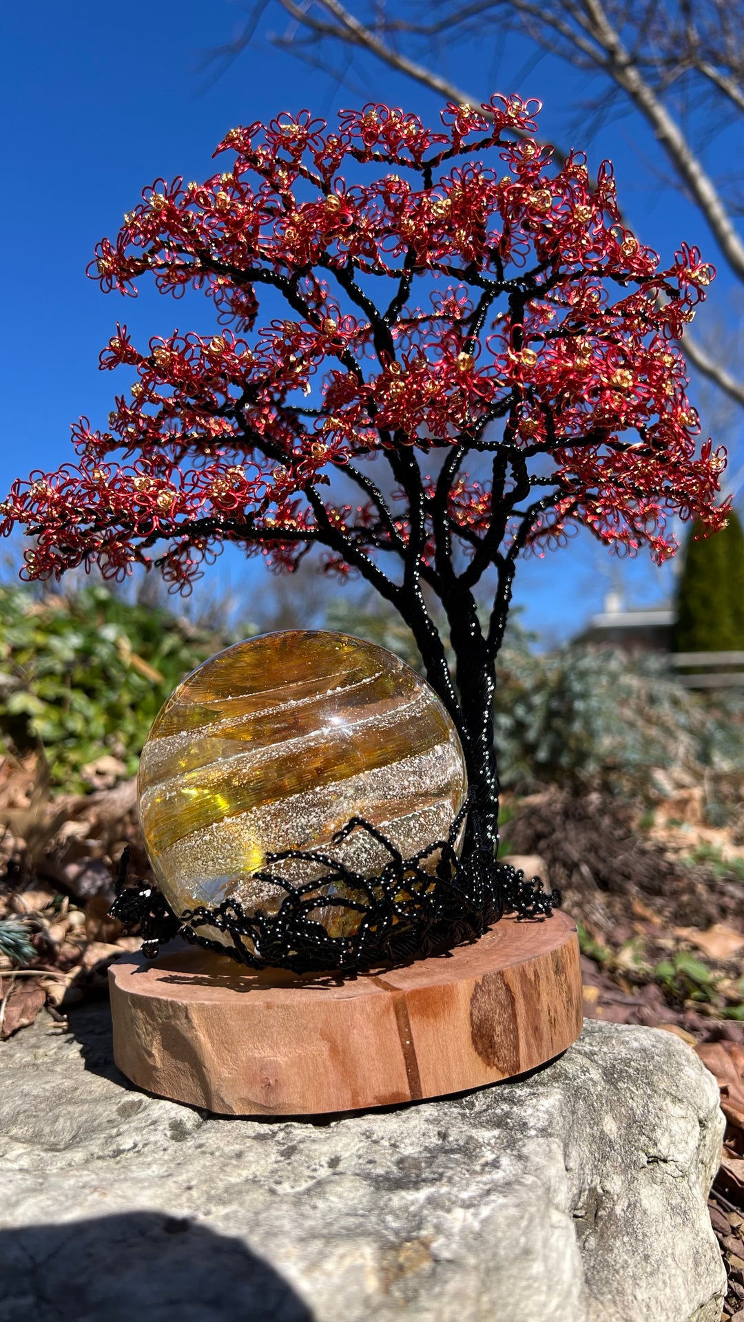 Close-up of Flamboyant Tree of Life wire sculpture with red blossoms and a gold Sparkle Orb on a wooden base outdoors