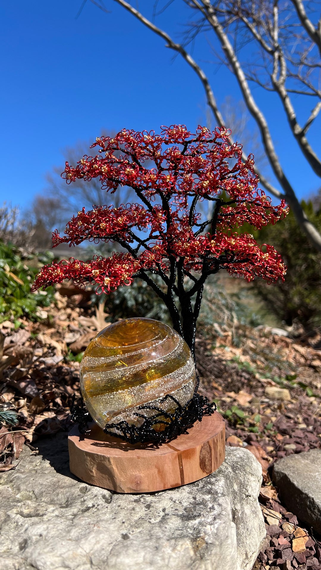 Flamboyant Tree of Life sculpture with red wire blossoms on a black trunk and a glass orb on a wooden base outdoors