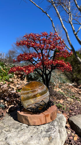 Flamboyant Tree of Life sculpture with red wire blossoms on a black trunk and a glass orb on a wooden base outdoors